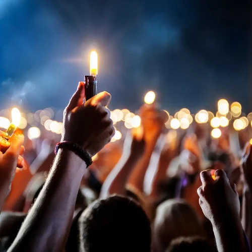 Hands holding lit lighters raised in the air at a nighttime gathering, with a crowd of people and glowing lights creating a vibrant, celebratory atmosphere.