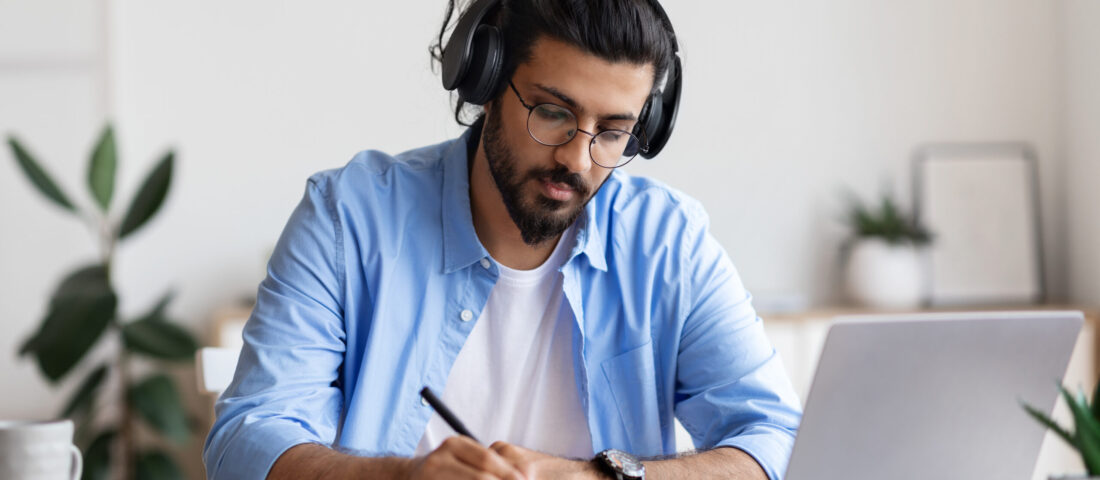 Distance Learning. Young Arab Guy In Headphones Studying With Laptop Computer At Home And Taking Notes To Notepad, Handsome Eastern Man Watching Webinar Or Having Online Training, Free Space